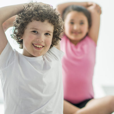 A multi-ethnic group of elementary age children are doing yoga at school. They are stretching their arms while sitting on their yoga mat.